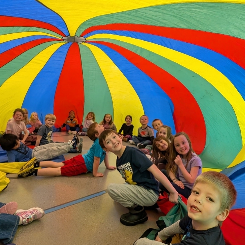 Students sit and smile under a large red, yellow, green, and blue parachute during PE class.