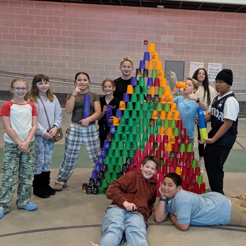 Students pose with a large colorful cup-stacking tower built in the gym during a PE activity.