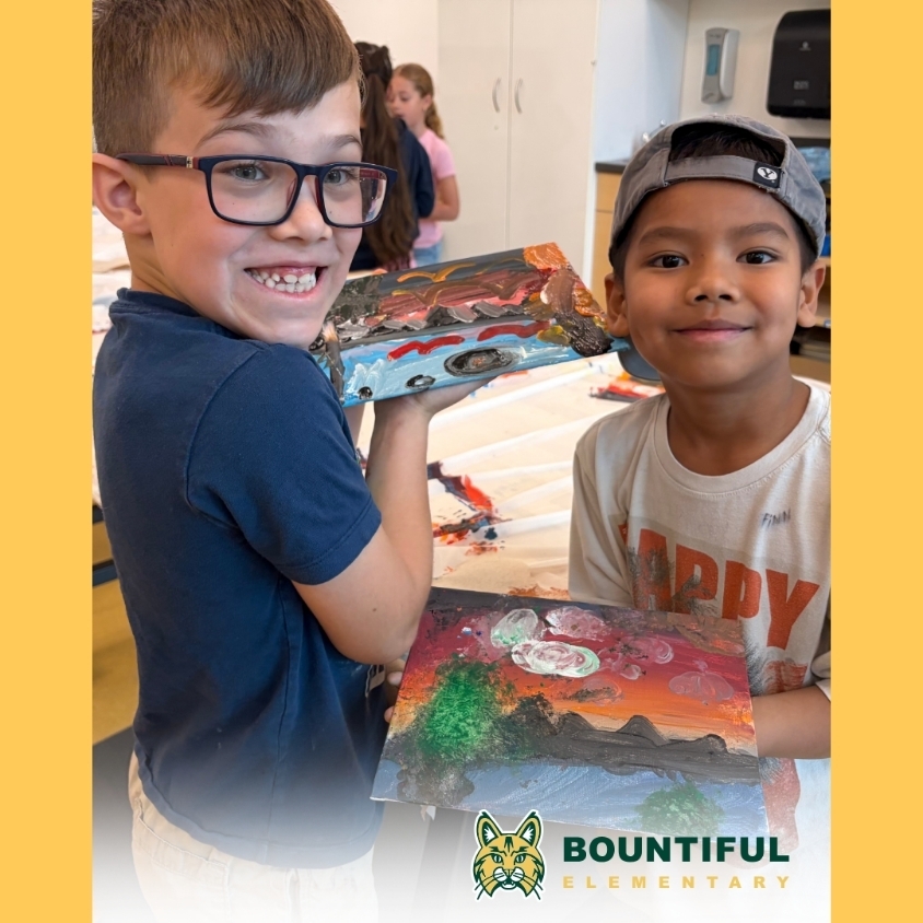 Two Bountiful Elementary students smile while holding colorful painted landscape projects in the art room.