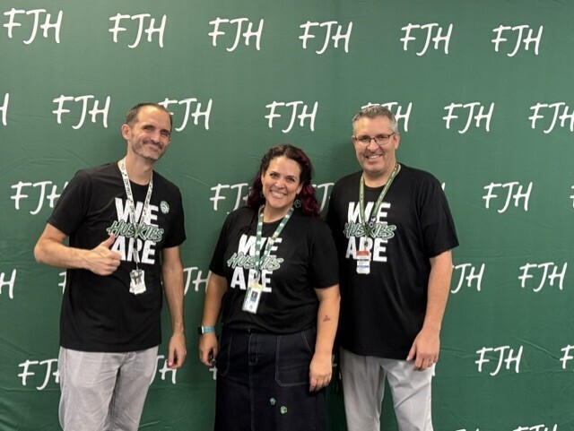 Three FJH school administrators smile for a photo wearing 'We Are Huskies' shirts, standing in front of a green FJH-patterned backdrop at Husky Camp.