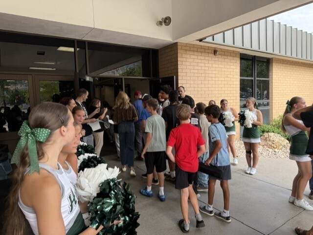 SBOs, Counselors, Administration, and Cheerleaders with green and white pom-poms welcome incoming 7th graders arriving for Husky Camp at the school entrance.