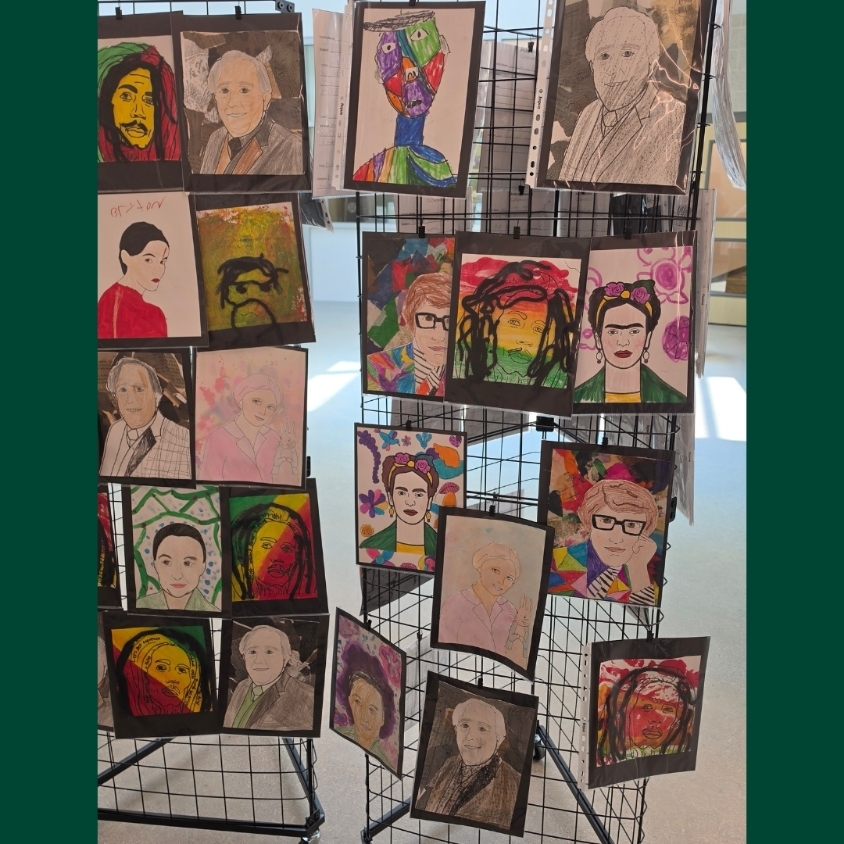 Multiple student portrait drawings and paintings are arranged on a freestanding hallway art display.