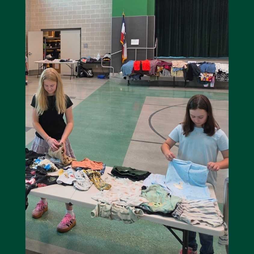 Two Hope Squad students fold and organize donated clothing at a table while additional sorted clothing is displayed on tables around the Bountiful Elementary gym.