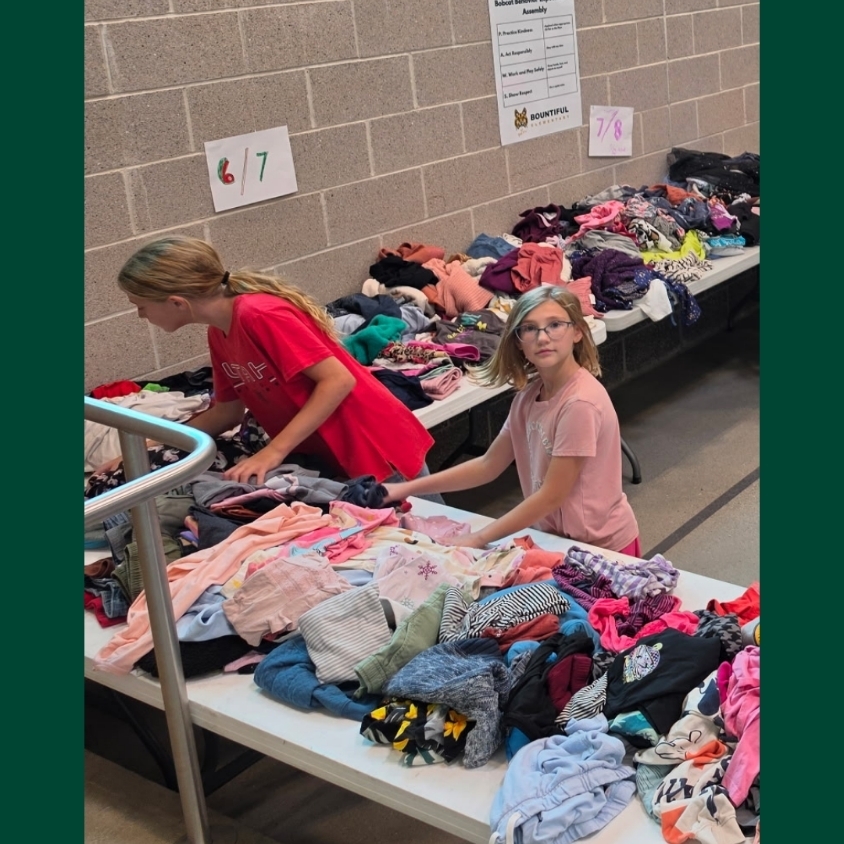 Two Hope Squad students sort donated children’s clothing at tables labeled by size, with large piles of shirts, pants, and other clothing ready to be organized for Back to School Night.