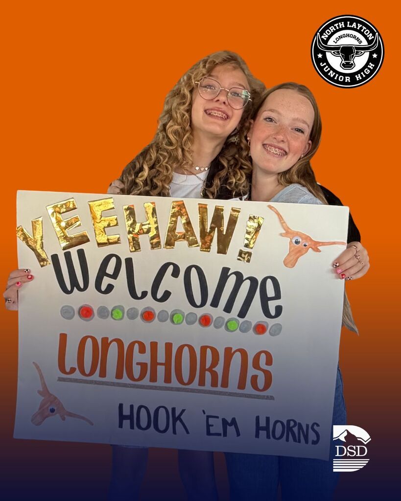 Two students stand outside holding a sign, greeting students as they enter the school