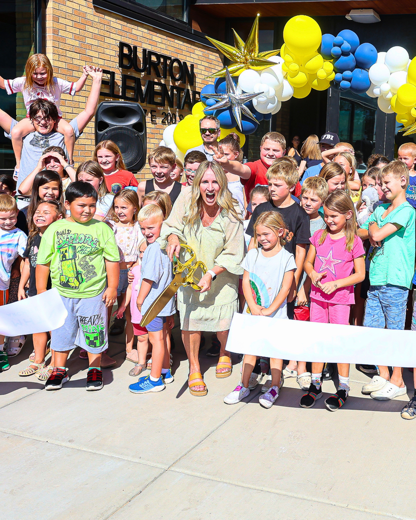 A bunch of students surrounding a principal who is cutting a large ribbon with gold scissors.