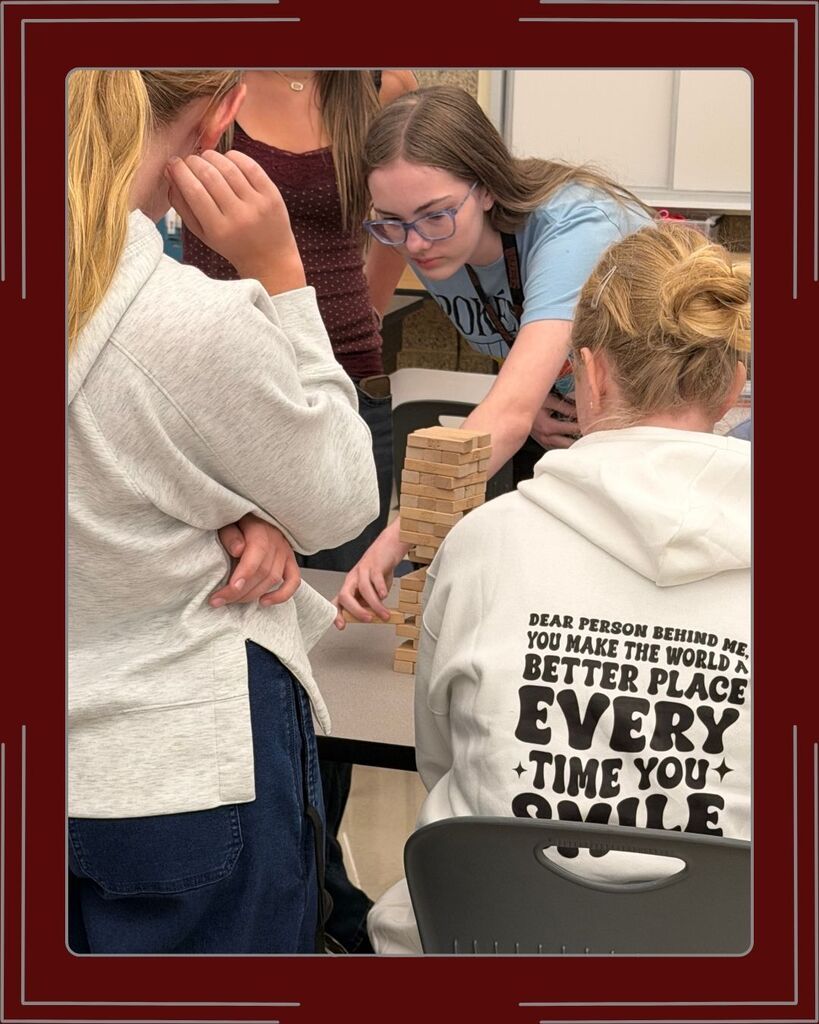 There are four girls from National Junior Honors Society in a classroom standing and sitting. The table has a tower of Jenga blocks; they are all very focused on the girl who is pulling a block out of the tower.