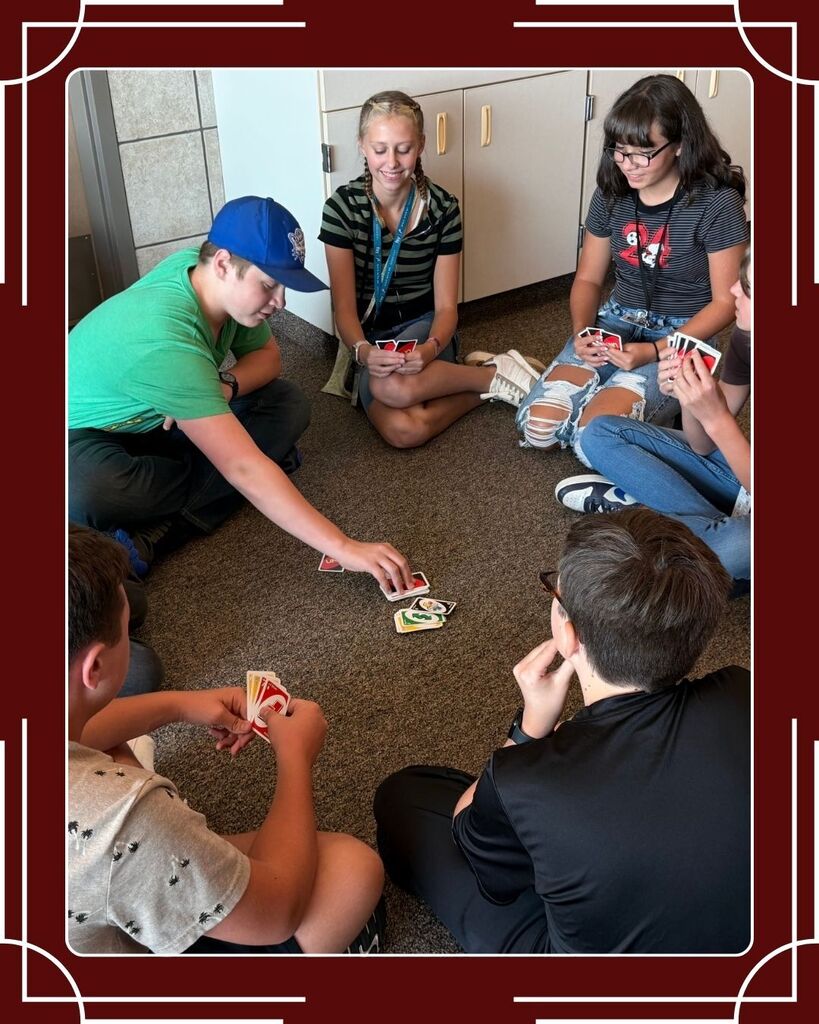 A group of National Junior Honors Society members are huddled around a game of UNO on the ground. One male student is drawing a card from the deck while the others are smiling and looking intrigued..