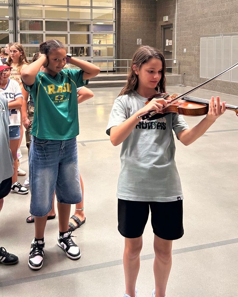 A student plays a violin during a hands-on music activity while classmates watch nearby. Other students can be seen waiting in the background for their turn to explore the instruments.