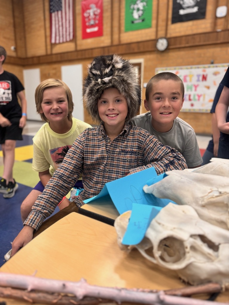 Three 4th grade boys sitting at a table in the cafeteria. One boy has a coon skin hat on and a plaid shirt.