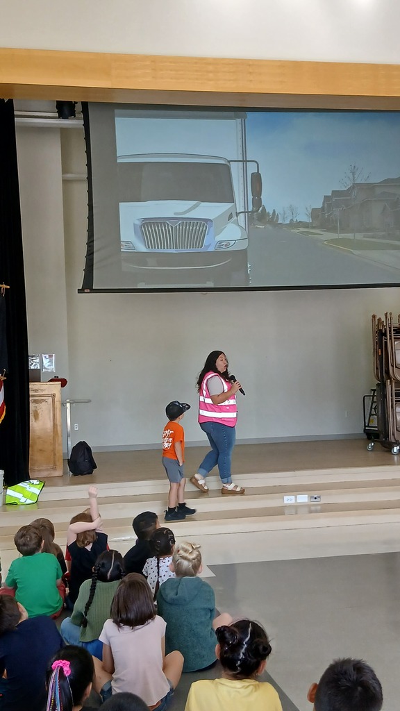 A presenter in a high-visibility pink vest talks into a microphone while a student volunteer stands beside her on stage, with seated students in the foreground looking on.