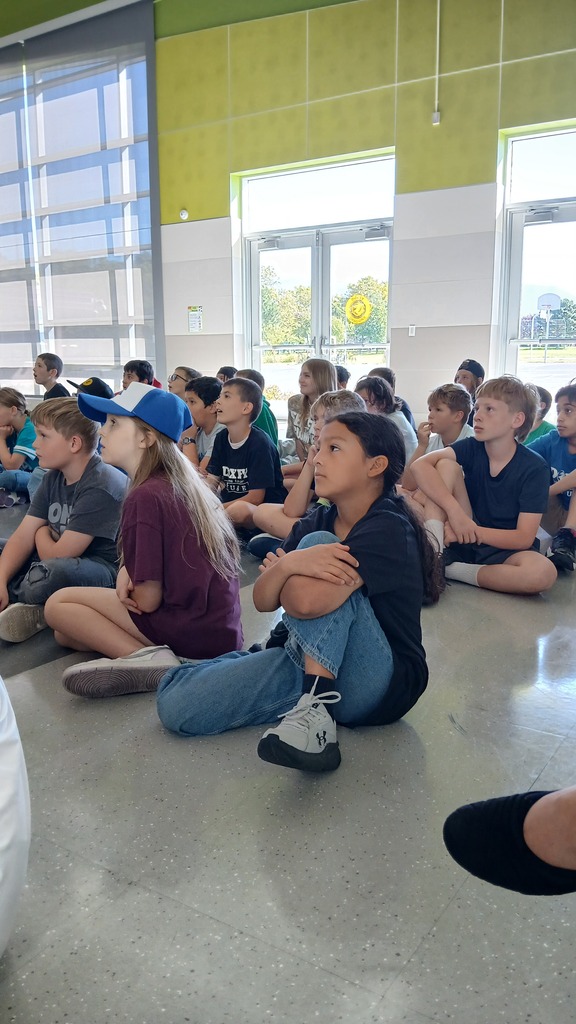 A group of elementary school students sitting cross-legged on the floor, looking attentively toward a assembly presentation off-camera.