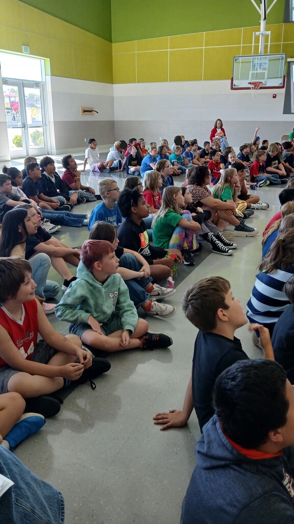 Students sitting on the floor in an auditorium facing a stage and projector screen that displays an instructional video about road safety.