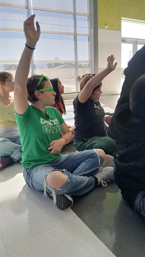 Elementary school students sitting cross-legged on the polished school floor with several children enthusiastically raising their hands during an assembly.
