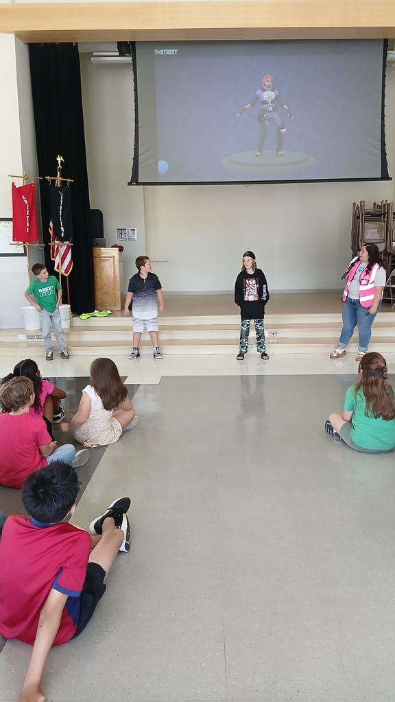 Three student volunteers stand on stage alongside a presenter in a high-visibility pink vest during an interactive presentation in front of an audience of seated students.