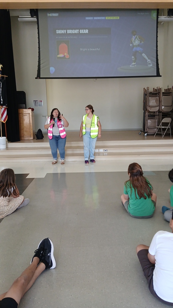 A presenter and a volunteer student in safety vests address students sitting on the floor, in front of a projection screen displaying a presentation about wearing "Shiny Bright Gear."