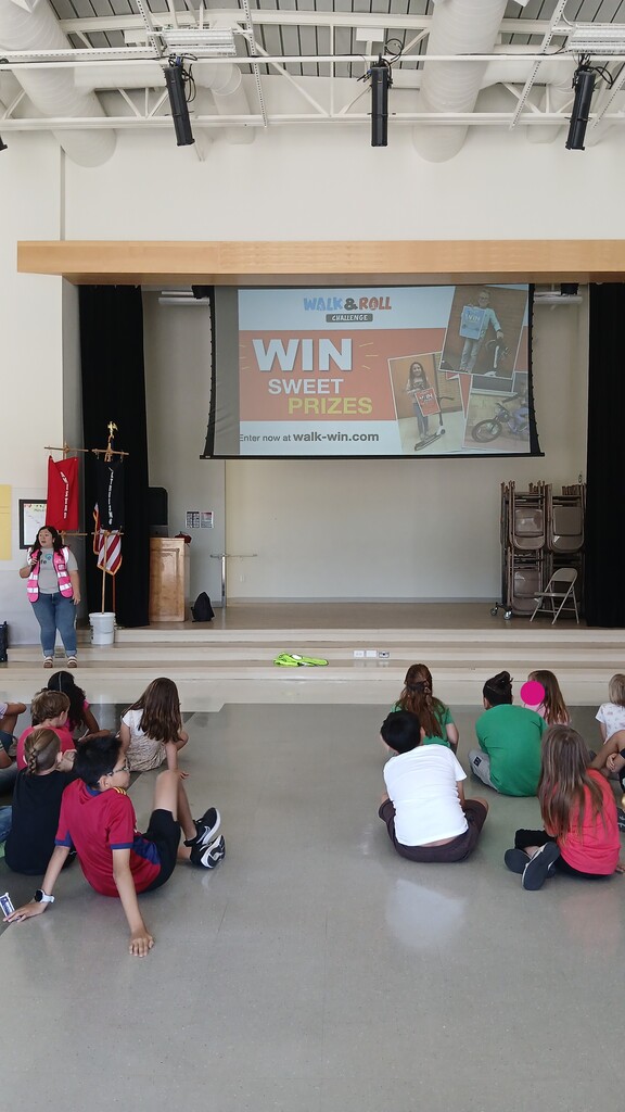 A presenter in a high-visibility pink vest stands on a stage next to a projector screen displaying "Walk & Roll Challenge - Win Sweet Prizes" while elementary students sit on the gymnasium floor watching.