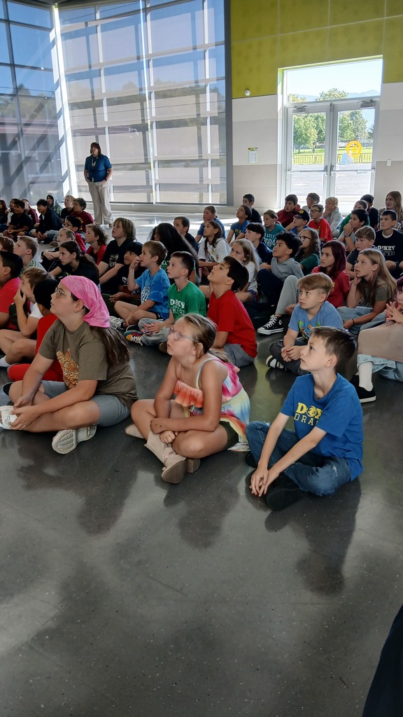 A wide view of students sitting on the floor in a bright, modern school multi-purpose room during an assembly presentation.