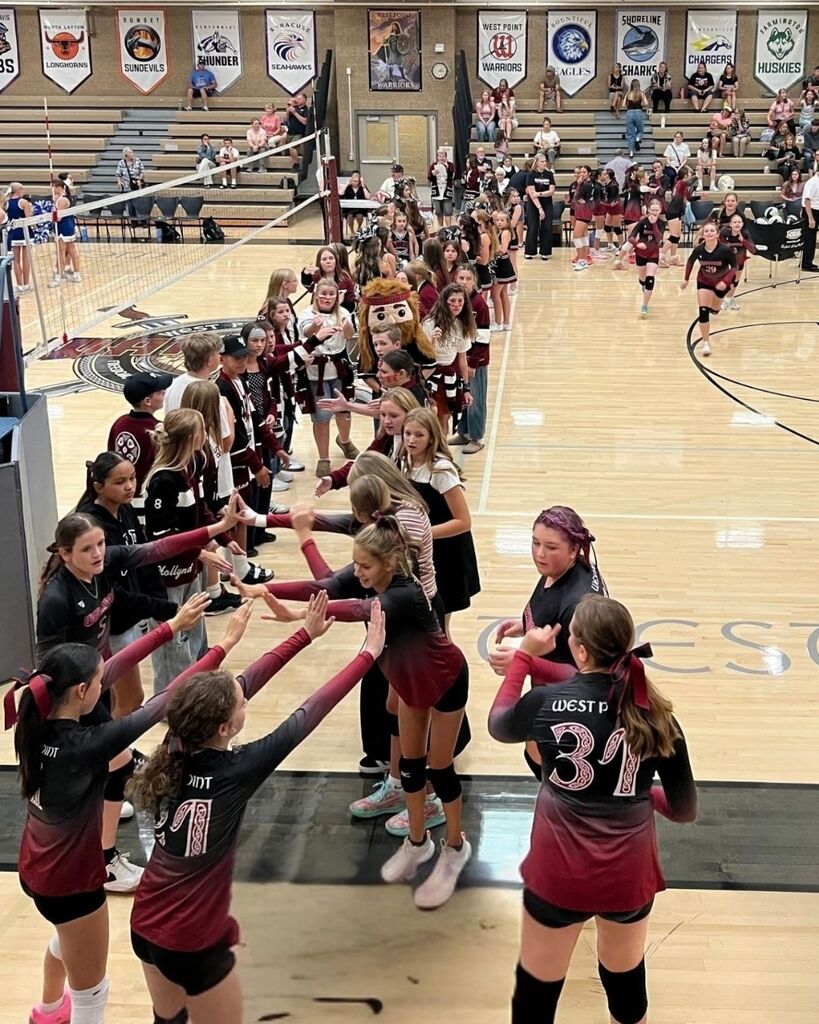Alt Text: West Point Junior High volleyball players in maroon-and-black uniforms form a high-five tunnel on the gym floor as teammates enter for a match. Students and spectators line the sides of the court and sit in the bleachers, while school banners from neighboring communities hang above. A West Point Warriors mascot stands among the crowd, creating a spirited game-day atmosphere.