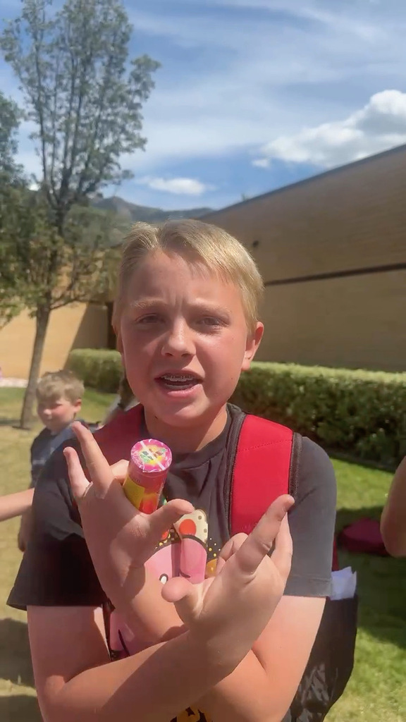 Blonde male student in black shirt and red backpack in front of his elementary school