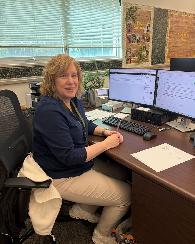 Female teacher dressed in a blue shirt and kaki pants sitting at her desk in front of two computer screens.