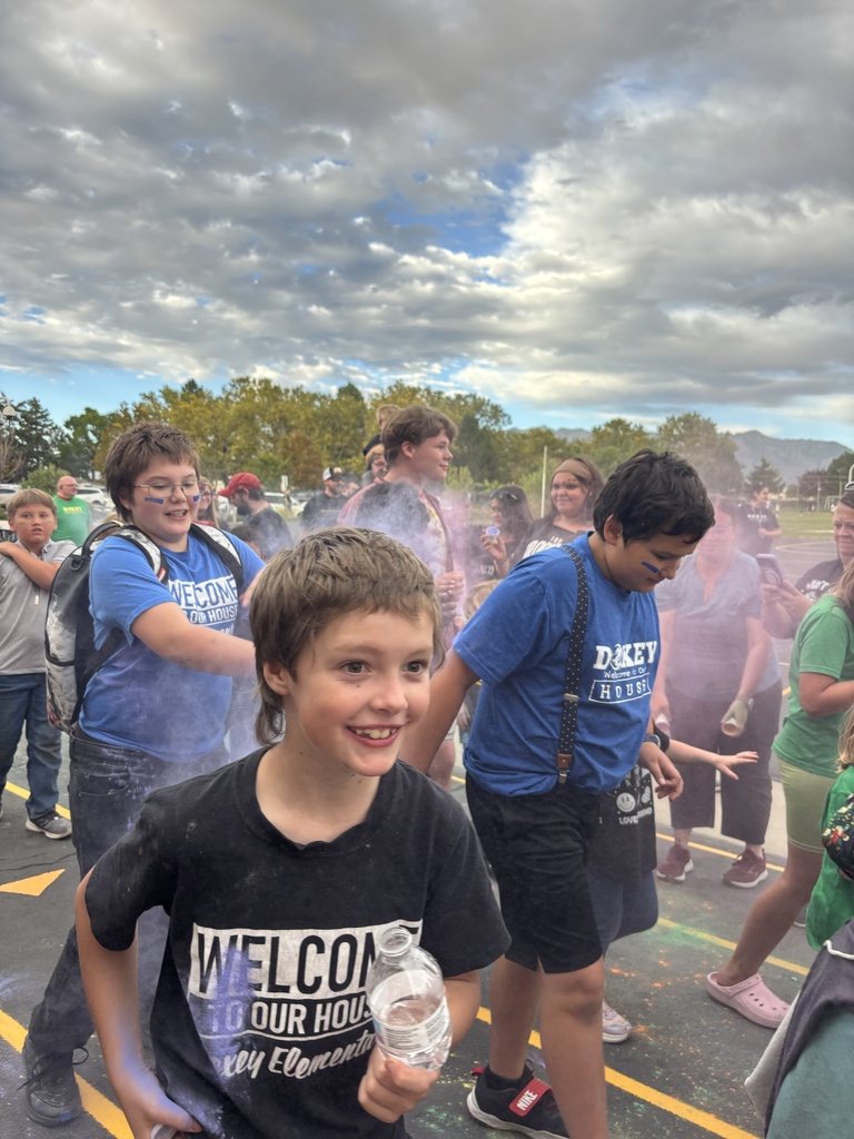 A smiling young boy with short brown hair walks outdoors in a school parking lot holding a water bottle, with other students in blue Doxey Elementary house shirts and clouds of colored chalk dust in the background under a cloudy sky.