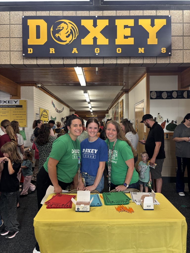 Three smiling staff members stand behind a yellow table set up in a school hallway beneath a large "DOXEY DRAGONS" sign, with colorful sorting papers and pencils laid out for the event.