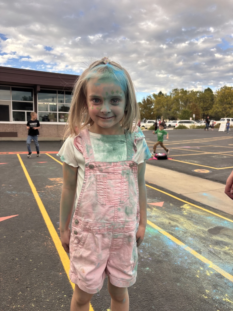 A young girl with her hair half-up stands outside, smiling warmly while covered in smudges of pink, blue, and green chalk dust on her face, white shirt, and pink overalls.