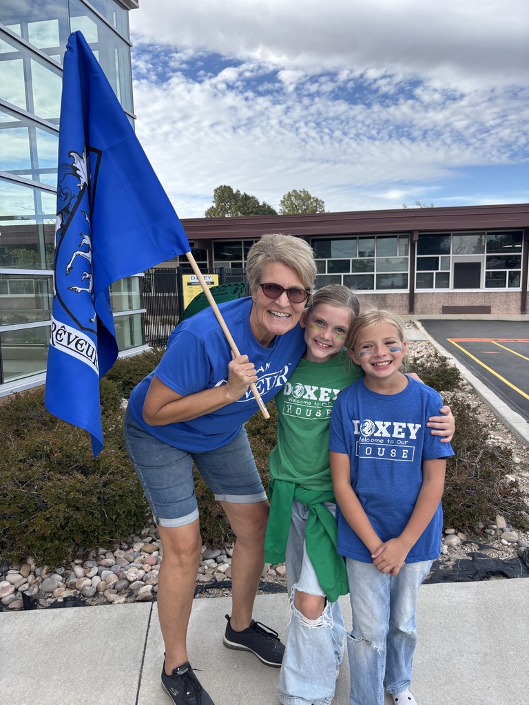 An adult holding a blue house flag smiles outdoors alongside two young girls wearing blue and green Doxey Elementary house shirts, standing on a sidewalk near a school building.