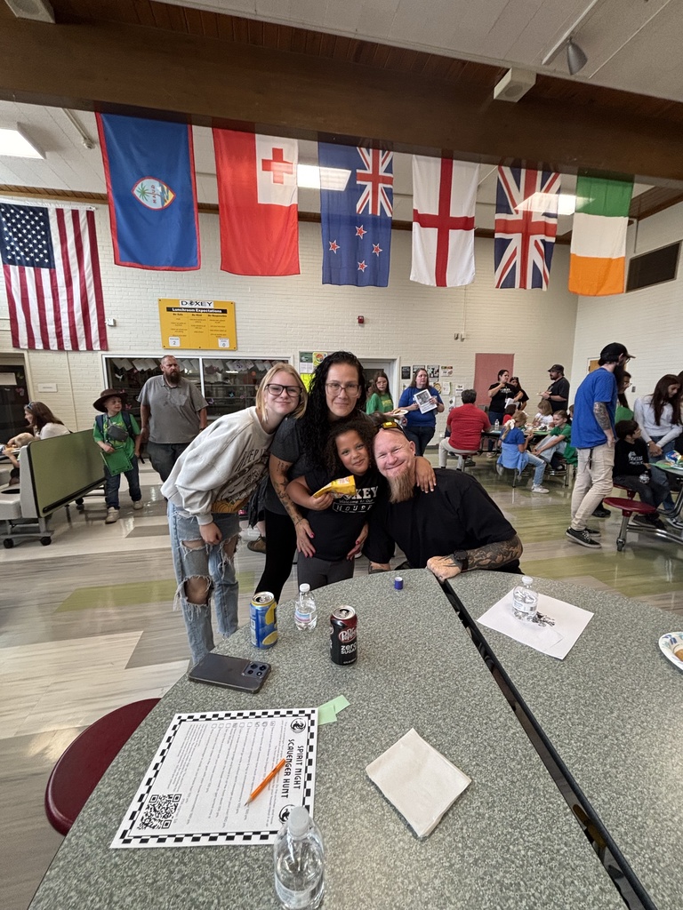 A family of four poses together at a table in a school cafeteria decorated with international flags hanging from the ceiling, smiling at the camera with drinks and a scavenger hunt paper on the table.