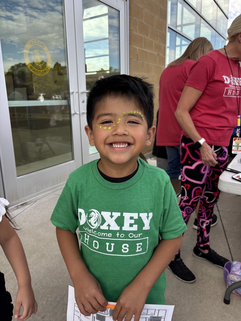 A young boy in a green "Doxey Welcome to Our House" shirt smiles brightly outdoors with yellow face paint markings on his cheeks, with school staff members visible in the background near a glass door.