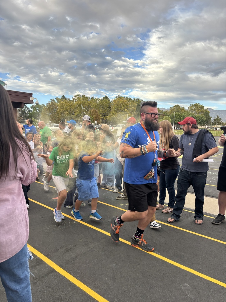 A group of adults and children run across a school parking lot during a chalk run event, kicking up clouds of yellow and green chalk powder while smiling and laughing under a dramatic cloudy sky.