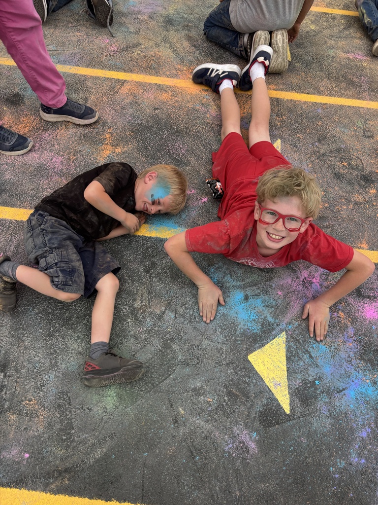 Two young boys play and laugh while lying on an asphalt school playground covered in scattered chalk dust; one boy wears a black shirt and the other wears a red shirt with red glasses.
