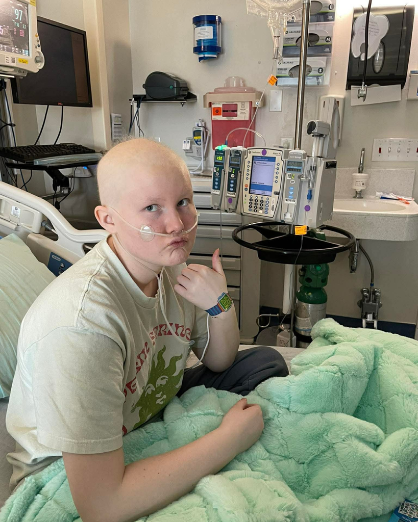 A student in a hospital bed with a blanket giving a thumbs up. 