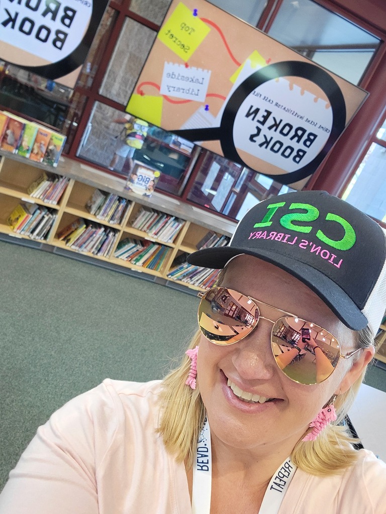 Mrs. Angie wearing a CSI Lions Library Hat and Broken Books display behind her.