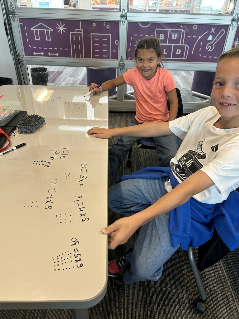 a boy and girl sitting around a desk with multiplication equations and dots written on the desk