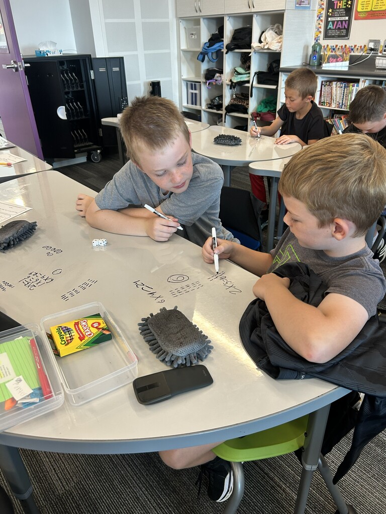 two boys using a dry erase marker on their desk to write a multiplication equation
