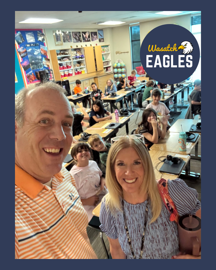 A classroom photo featuring two adults standing at the front of a fourth-grade classroom while students work at desks in the background. Bookshelves, classroom materials, and educational posters are visible around the room. A Wasatch Eagles logo is displayed in the upper-right corner of the image. Students are seated throughout the classroom, engaged in learning activities.