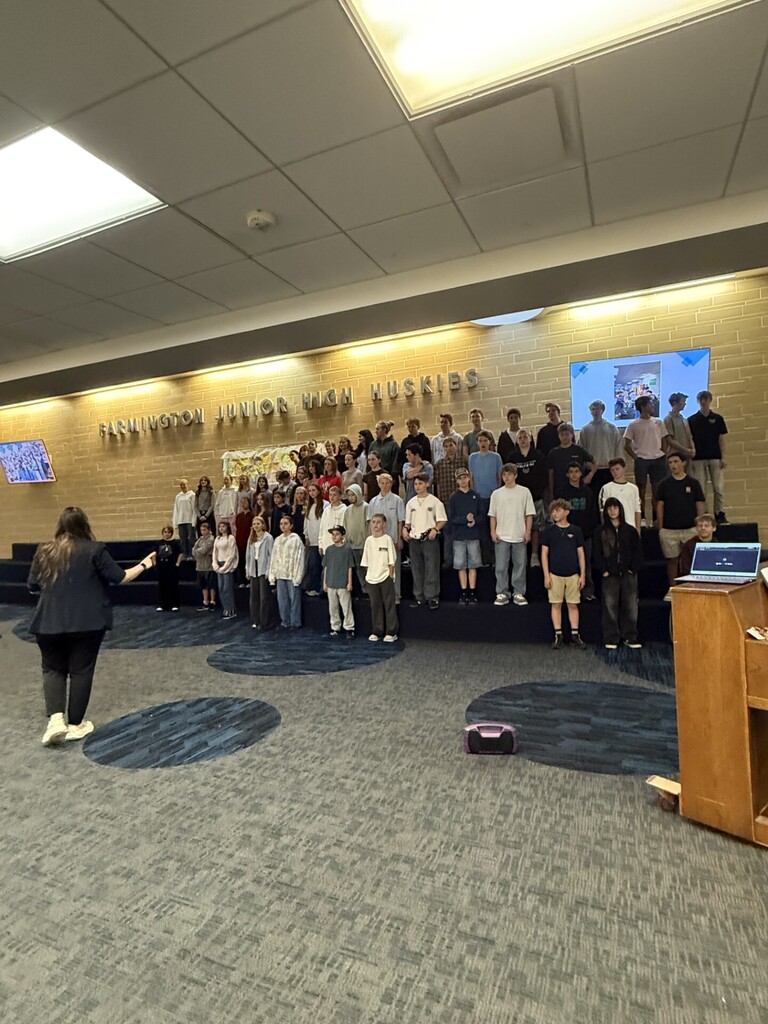A choir director leads a large group of junior high students standing on risers in the school commons, in front of a "Farmington Junior High Huskies" sign.