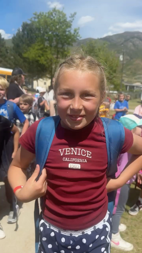 Blonde girl in red shirt and blue backpack standing in front of school