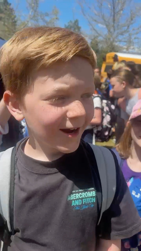 Red headed boy in black shirt standing in front of school with many students