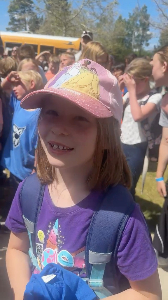 Female student in pink hat and purple shirt in blue backpack standing in front of school with other students