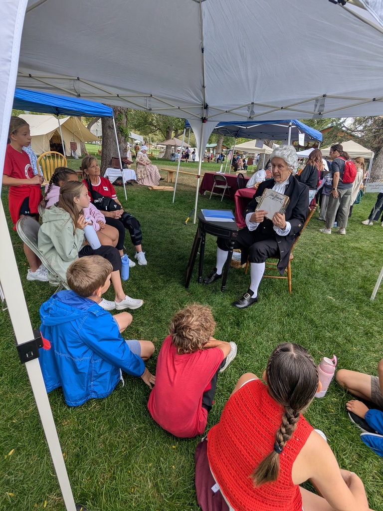 Students sitting on the ground listening to a gentleman in colonial clothing. 