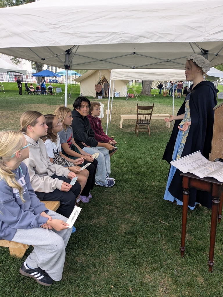 A group of students sitting down, listening to a lady dressed in colonial clothing.
