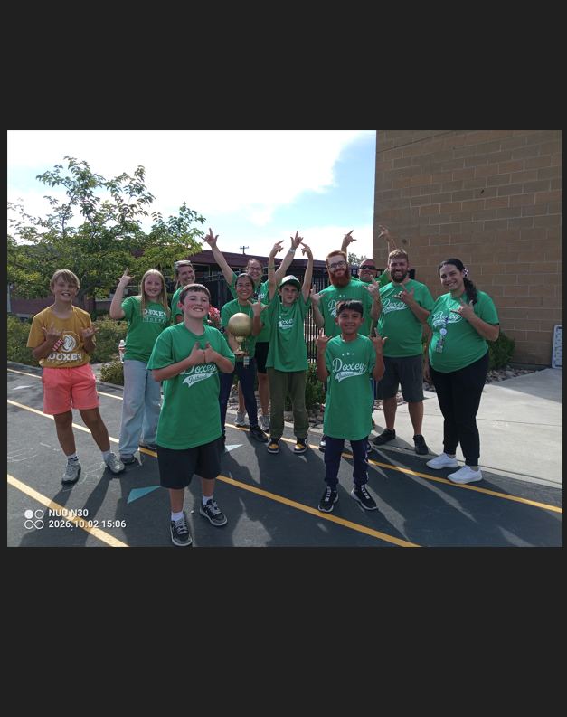 A group of 12 people, a mix of adults and children, are celebrating and cheering outdoors on a paved area next to a brick building. All are wearing matching bright green t-shirts with "Doxey" printed on them. The person in the front center is holding up a small, shiny, gold-colored trophy cup. Several participants are raising their arms in the air. The photo has a date stamp in the bottom left corner.
