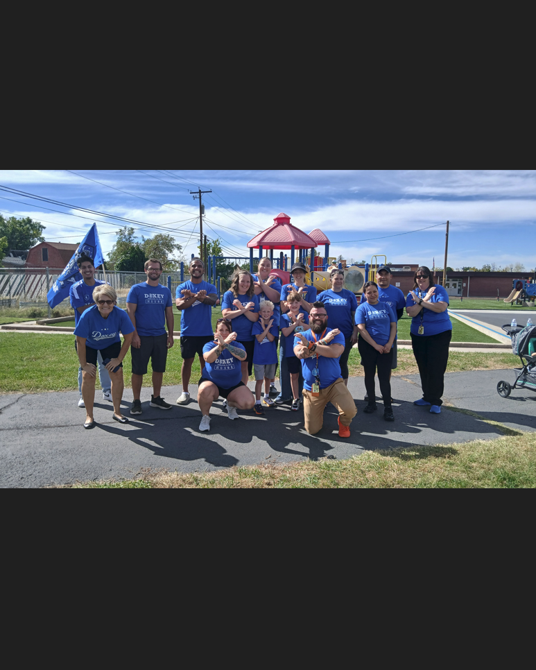 A group of 13 people, mostly adults and some children, are posing for a group photo on a paved path and grass. All are wearing matching royal blue t-shirts with "Doxey" on them. A blue flag with a logo stands on a pole to the left, and a red-roofed playground structure is in the background. Some people are striking a celebratory or "X" pose with their arms.