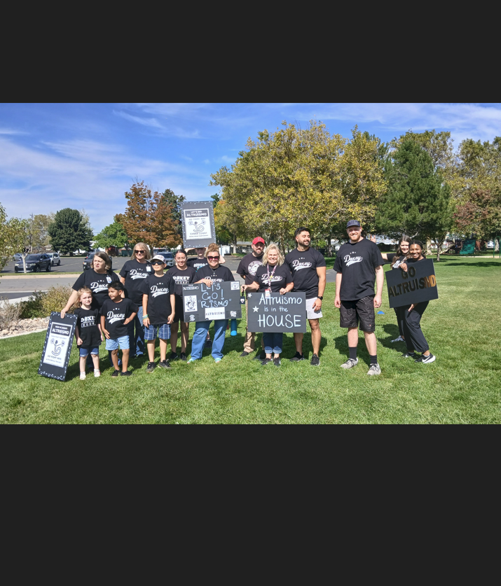 A large group of about 18 people, primarily wearing black t-shirts with "Doxey" printed in white, stand for a group photo on a grassy field. Several people in the front hold small signs. Two people in the back hold a larger sign that reads "Altruismo is in the HOUSE". Trees and a street are visible in the background under a blue sky.