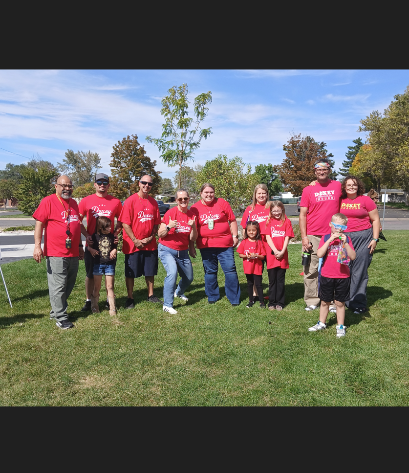 A large group of approximately 15 people, including adults and children, are gathered on a grassy area. Most are wearing matching red t-shirts with "Doxey" on them. They are posing for a group photo next to a whiteboard on an easel, with trees and a street in the background. A person in a blue shirt stands to the left, outside the main group.