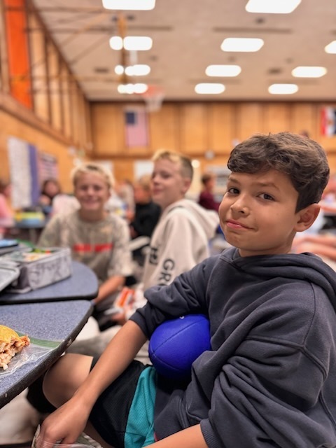 5th grade boy in black hoodie and holds his blue football sits in the cafeteria with his friends