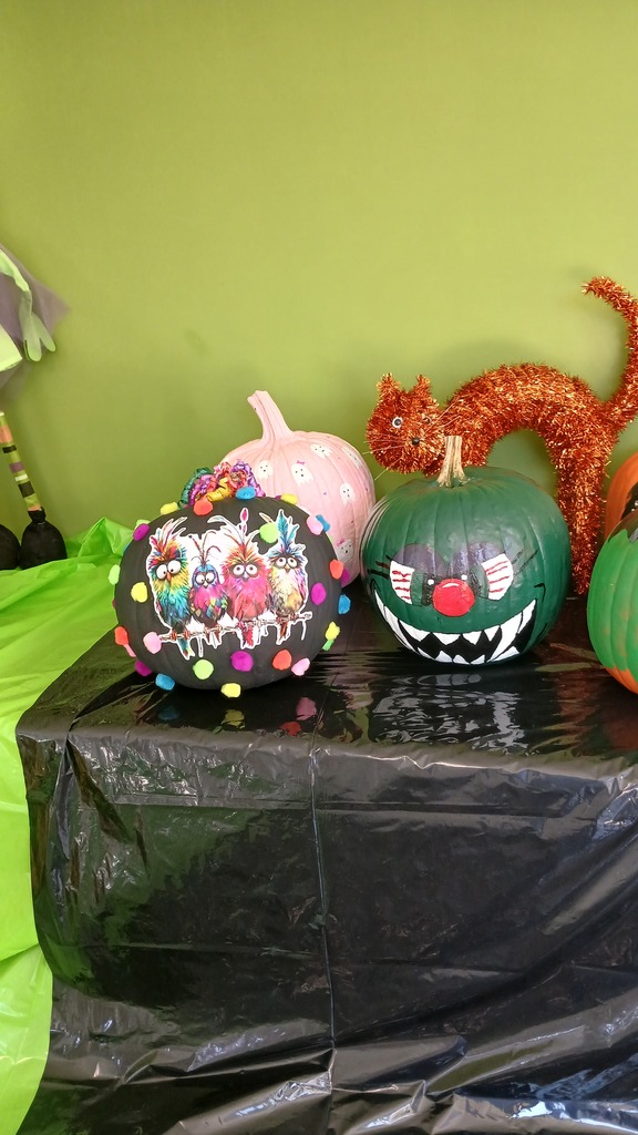 A vibrant cluster of painted pumpkins on a black table covering, showcasing a dark pumpkin decorated with colorful whimsical owls and pom-poms, a pink ghost pumpkin, and a green monster face with a red nose
